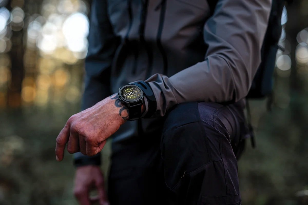 Närbild på en person i skogen med en robust taktisk Garmin smartwatch på handleden, iklädd mörka friluftskläder och med armen vilande mot knät.